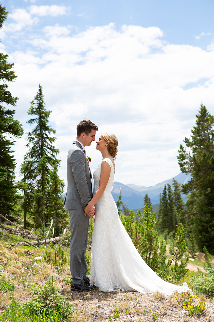 Antasia and Jacob hold hands in a pine forest with the mountains behind during their wedding in Colorado.