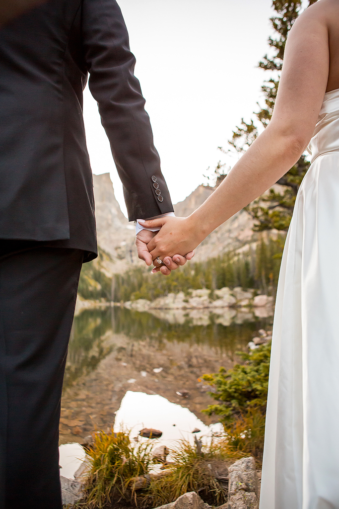 Close up hands and wedding ring with an alpine lake and mountain peak in the background.