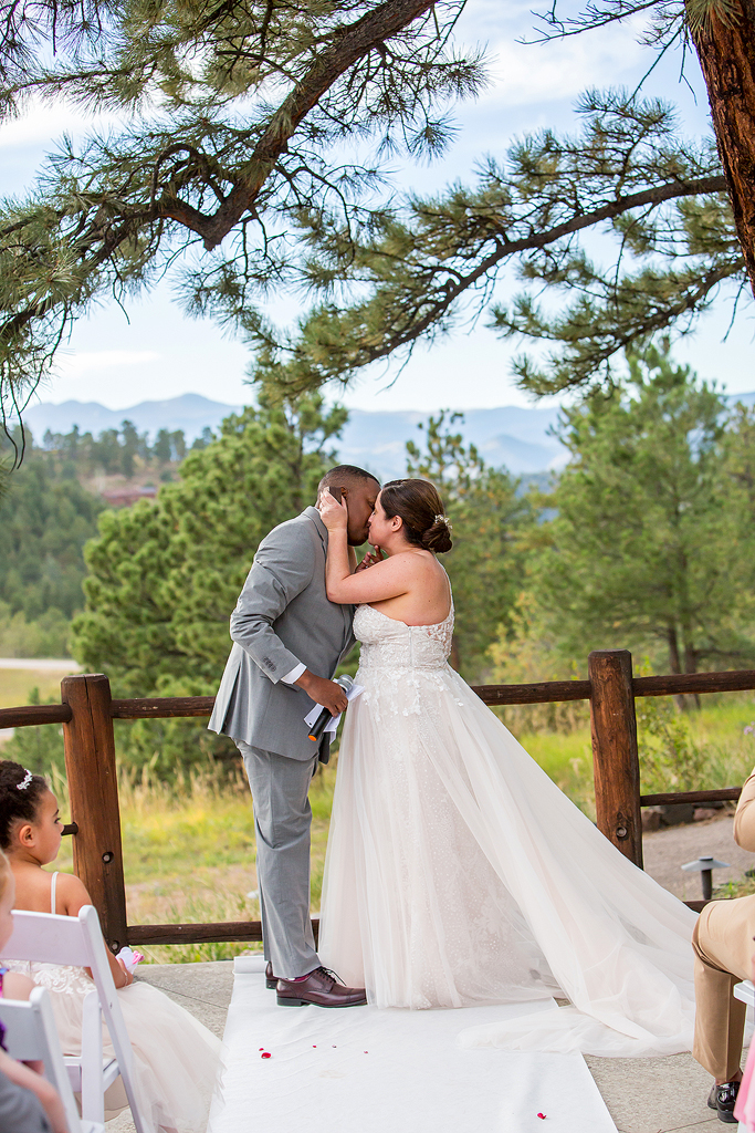Bride and groom kiss after their ceremony under a large pine tree with mountain behind them.