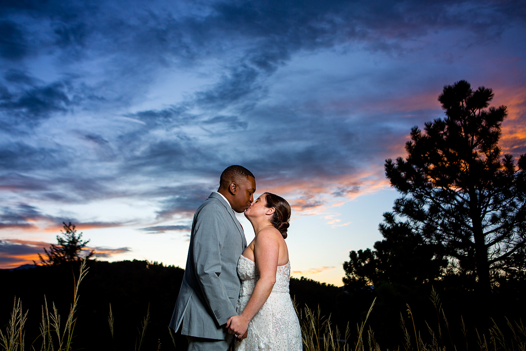 Bride and groom kiss in front of a colorful sunset near Evergreen, Colorado.