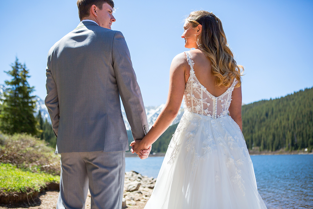 Bride and groom hold hands in front of an alpine lake in Colorado.