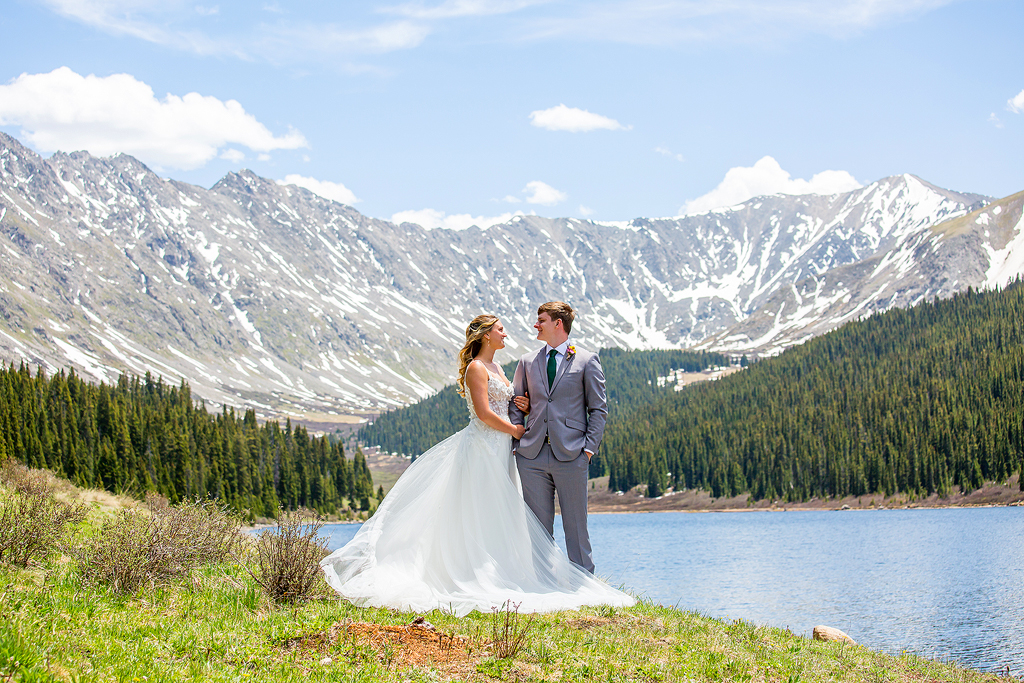 Wide view of snow capped mountains, forest, and alpine lake in Colorado with bride and groom in the foreground.