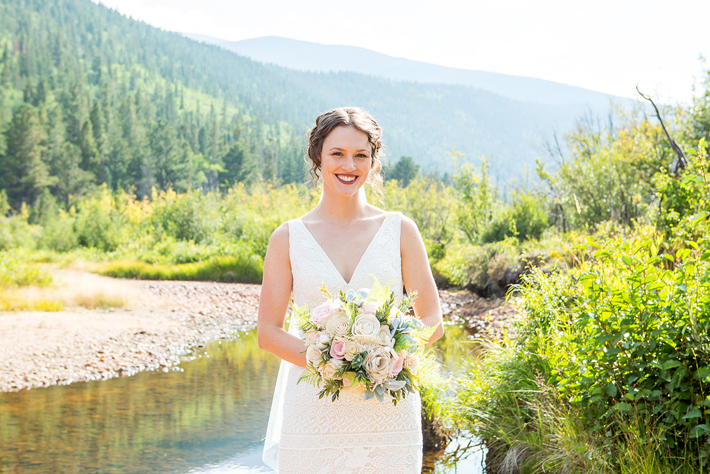 Bride holding her bouquet in front of a pine forest and creek in Colorado.