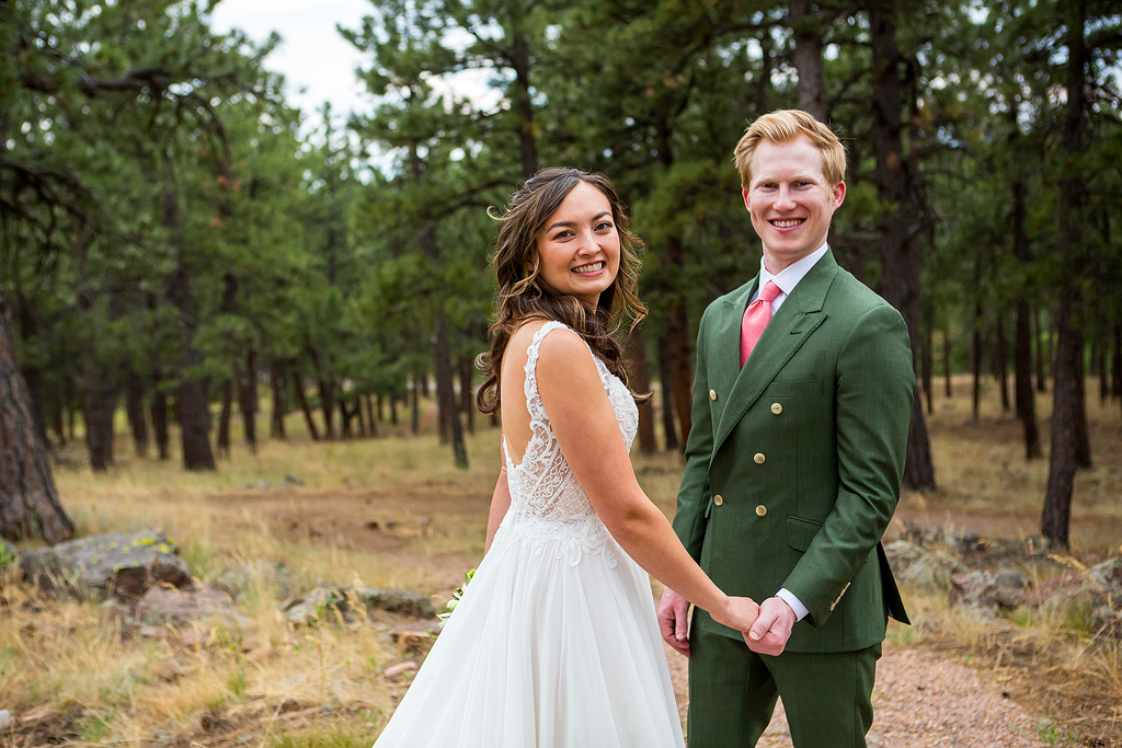 Bride and groom hold hands and smile surrounded by pine trees.