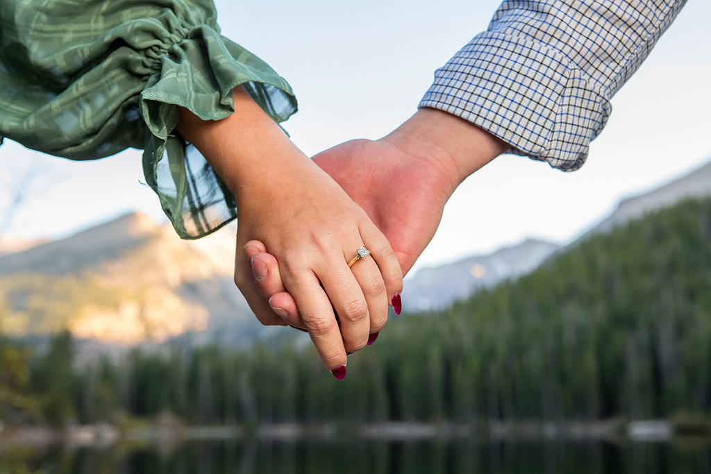 Close up of engagement ring and couple holding hands with mountains and lake in the background
