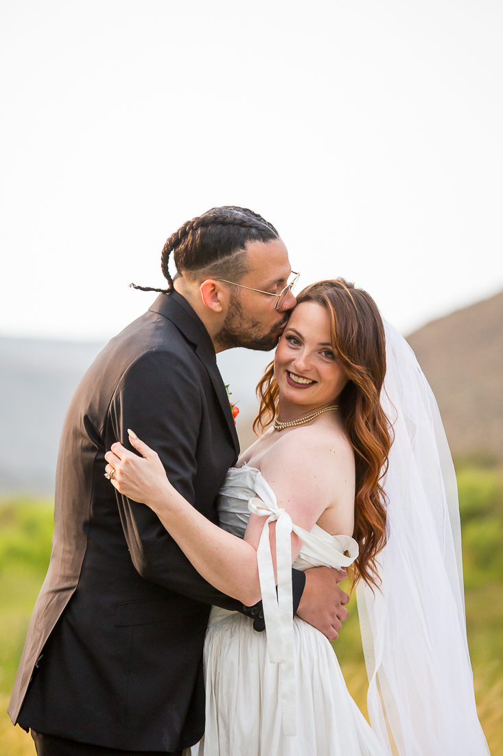 Bride smiles at the camera as groom kisses her on the temple in a field in Colorado.